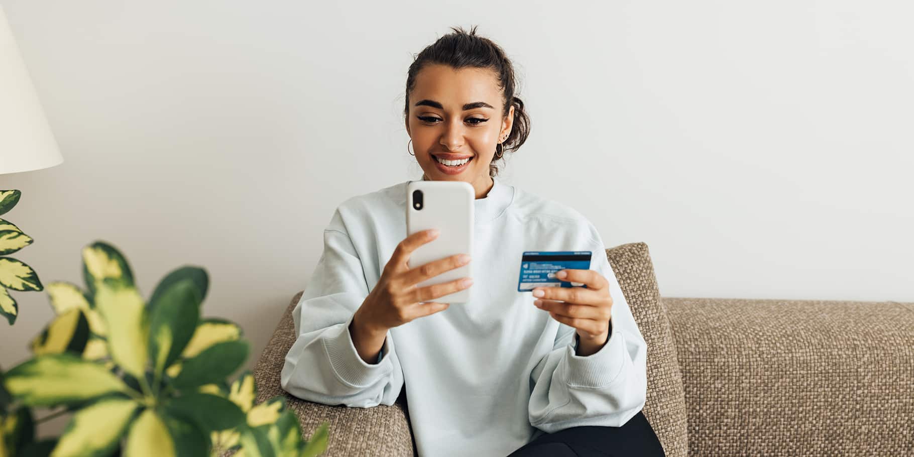 A young woman sitting on a couch while holding a credit card and a smart phone.