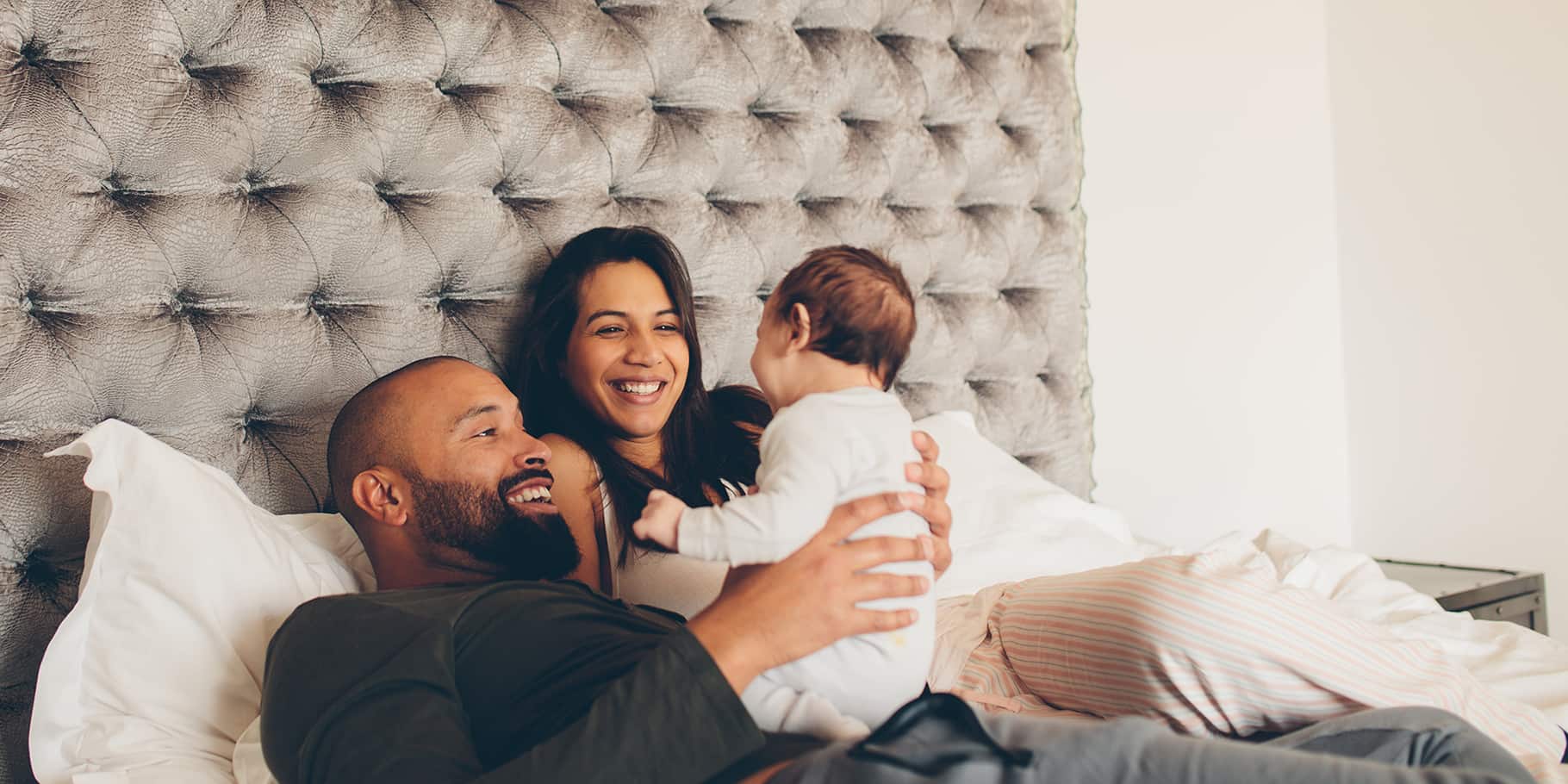 A smiling father and mother with their baby on their bed.