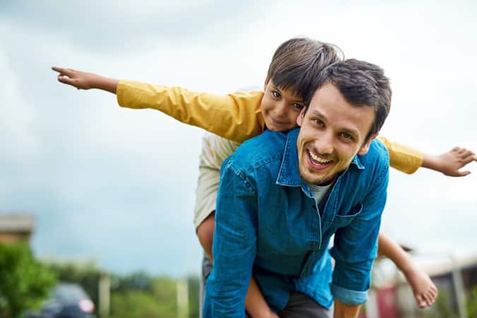 An adult male carrying a young boy with outstreatched arms on his back.