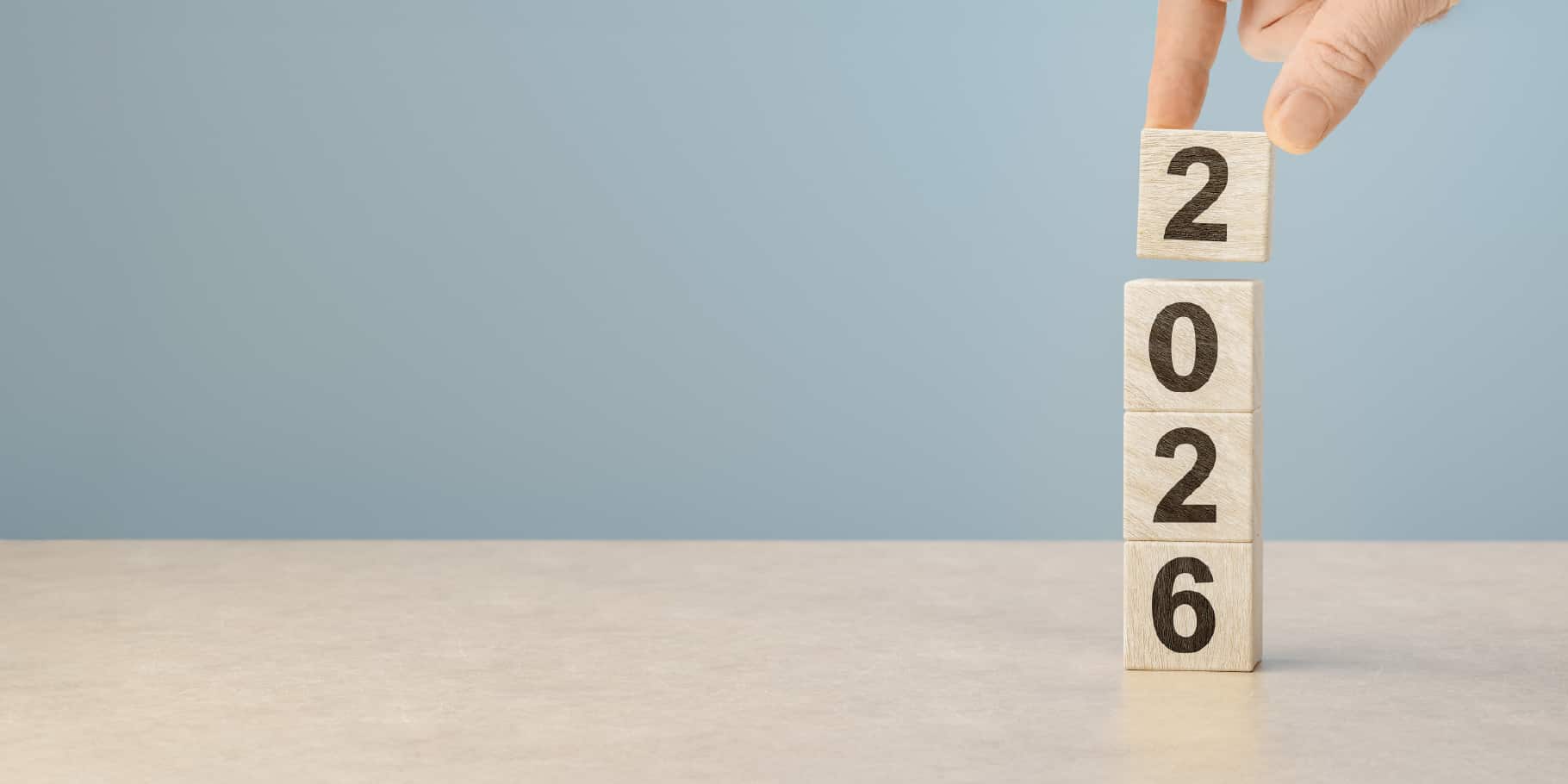 Wooden blocks stacked to display the year 2026, with a hand placing the final block on top against a soft blue background.