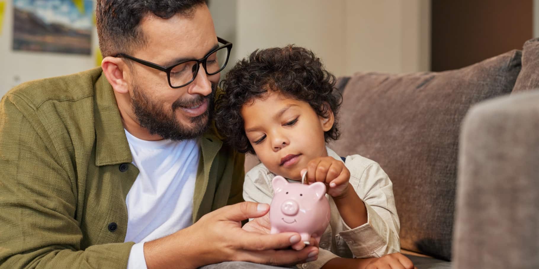 A father and young son sit together while the child puts a coin into a piggy bank.